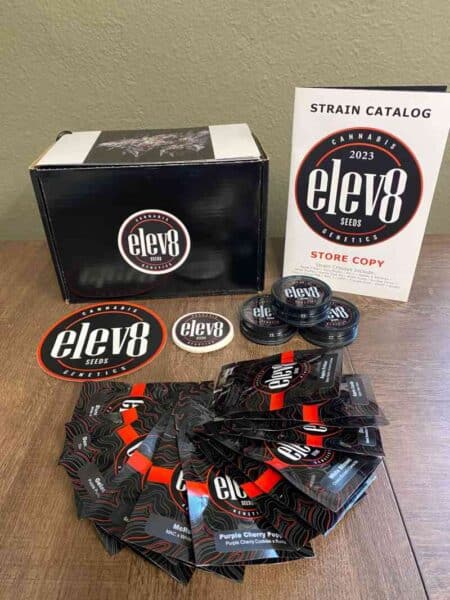 A collection of Elev8 Seeds promotional materials, including seed packages, stickers, a plastic container, and a 2023 strain catalog featuring the highly acclaimed elev8 Bakers Dozen, are displayed on a wooden surface.