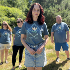 Four people standing outdoors on grass, one woman in the foreground wearing a denim skirt and a Cultivate Freedom T-Shirt, with three others in similar outfits in the background. Trees and sunlight fill the backdrop as they celebrate Pride Month, sparking the rainbow with their joyful presence.